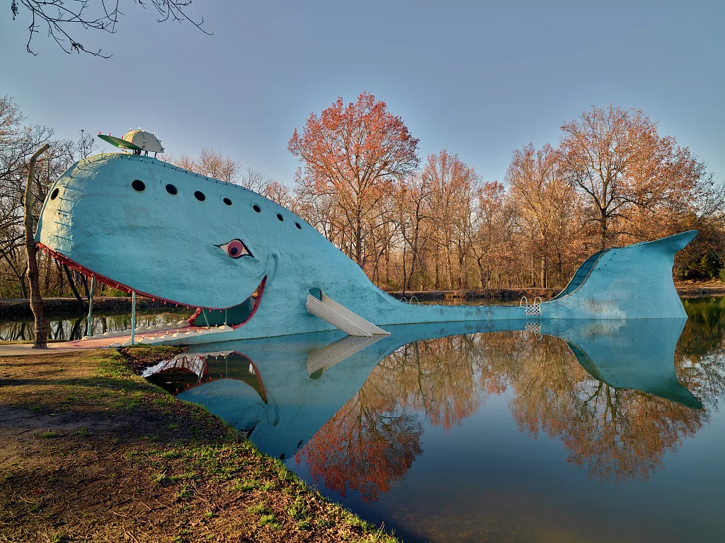 A bright blue concrete whale stretches across a pond, reflected in the water beneath autumn trees.